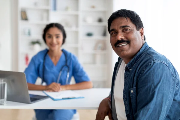 Portrait Of Happy Mature Indian Male Patient Sitting At Doctors Office, Smiling Man Satisfied After Successful Medical Treatment, Looking At Camera While Female Therapist In Uniform Posing At Desk