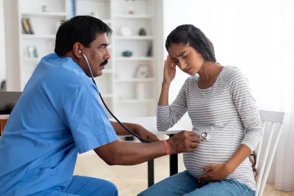 Prenatal Care. Indian Obstetrician Doctor Doing Check Up To Sick Pregnant Woman In Maternity Clinic, Eastern Male Gynecologist Examining Belly With Stethoscope, Listening Babys Heartbeat