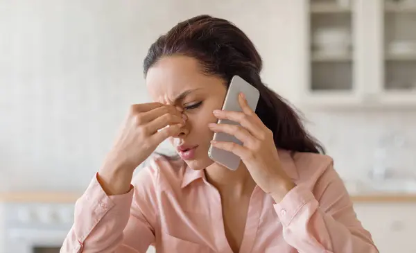 怒っている女性は 携帯電話で話し 鼻の橋をマッサージし 悪いニュース 圧力に苦しんでキッチンに座っています 自宅での問題 ストレス コミュニケーション — ストック写真