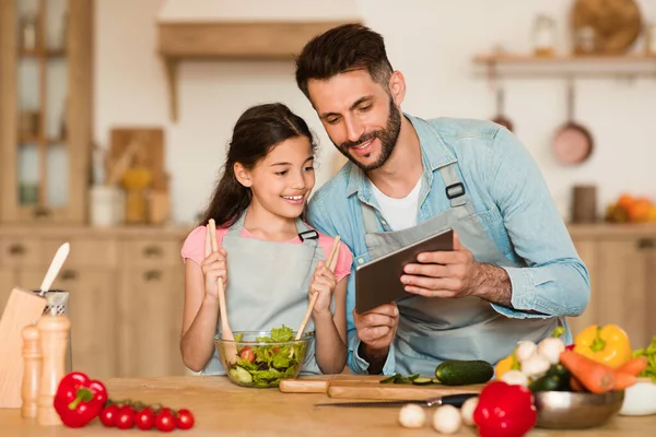Happy european dad and his daughter cooking in the kitchen, smiling man using digital tablet, watching food video blog or reading recipe online on culinary website