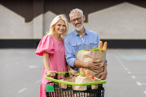 Portrait of happy elderly husband and wife shopping together at hypermarket, posing outdoors at mall parking spot. Cheerful retired caucasian couple holding packages with grocery. Healthy lifestyle