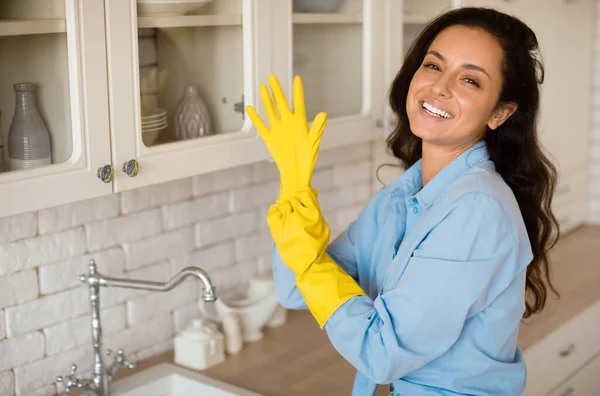 Portrait of joyful woman putting on rubber gloves, ready to tidy house or washing dishes, lady smiling at camera, standing in kitchen, free space. Cleaning service worker preparing to do housework