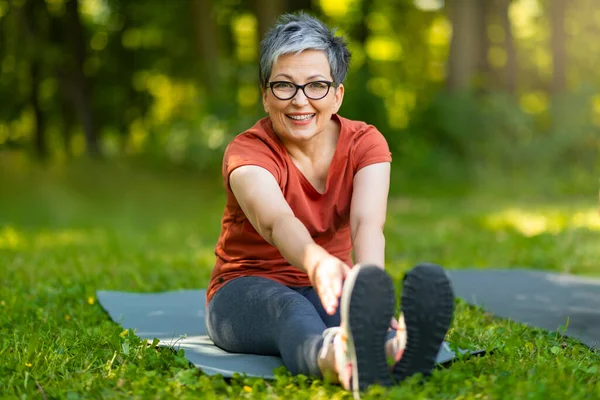 Beautiful Happy Senior Woman Training Outdoors, Smiling Older Lady Stretching Leg Muscles While Sitting On Fitness Mat On Lawn In Park, Sporty Mature Female Looking At Camera, Copy Space