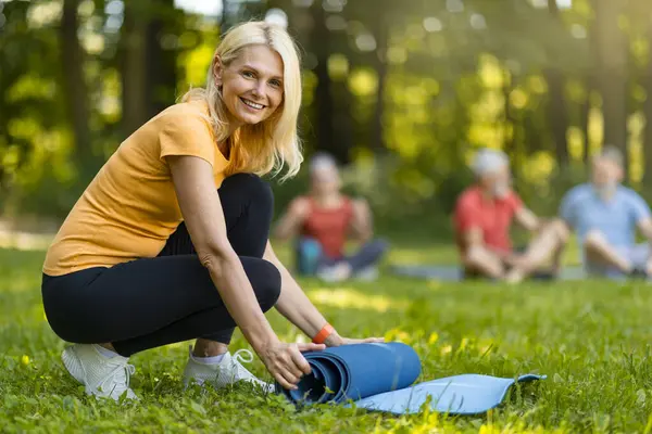 Mutlu Olgun Bayan Yuvarlanan Yoga Mat Spor antrenmanından sonra dışarıda, aktif giyimde gülümseyen yaşlı kadın grup spor sonrası dinlenme dersi, dışarıda antrenmanların ve sağlıklı yaşam tarzının tadını çıkarıyor.