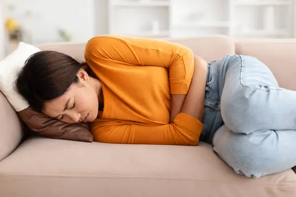 中国女人痛苦不堪地躺在沙发上 抱着肚子 经期肚子痛 特写镜头 有病的亚洲小姐肚子痛 食物中毒 — 图库照片