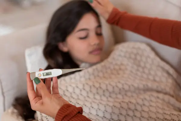 熱がベッドに横たわる病気の少女は 高温を示す温度計を保持している間 屋内装置に選択的な焦点を当てています 母親は病気の娘を大切にしています インフルエンザの治療 — ストック写真