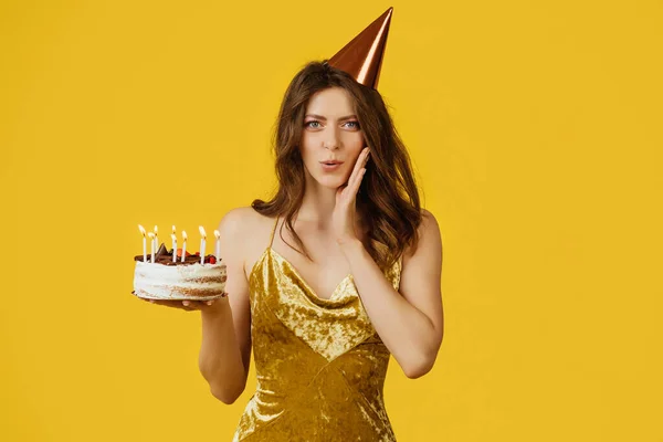 Happy young european woman in birthday hat making wish, holding cake with candles, isolated on yellow studio background. Dream, ad and offer, celebration birthday party