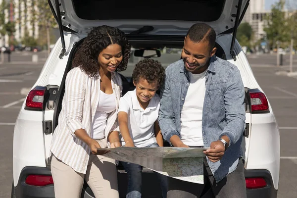 Happy black family father mother and preteen son going car trip together at weekend, standing on the street next to white automobile, checking map, looking for destination to travel