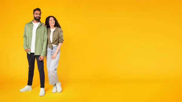 Sale offer. Middle eastern couple posing looking aside at empty space for text, woman leaning on mans shoulder, standing on yellow studio background, wearing casual clothes. Panorama, full length