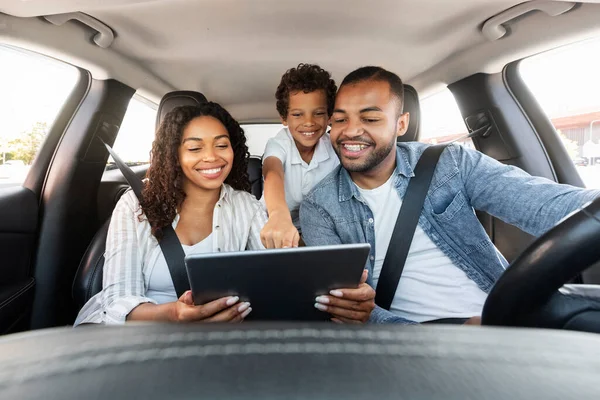 Positive cheerful black family have car ride, using digital tablet, checking map navigation while travelling. Cute african american kid boy point at pad screen in his mother hands, father driving auto