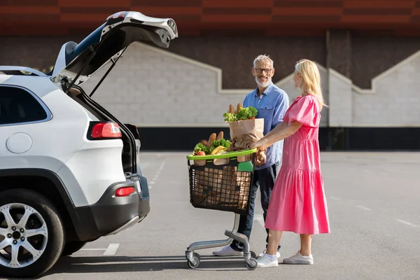 Loving attractive caucasian elderly couple standing next to white car with open trunk, senior man and woman husband and wife load grocery packages into auto, parking area background, full length