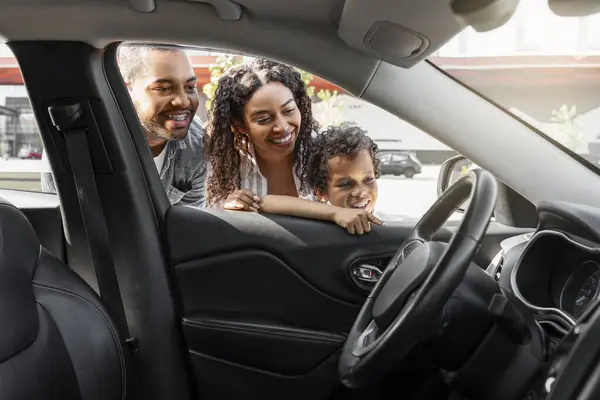 Friendly african american family choosing new car at dealership center parking spot. Excited black mom, dad and preteen child son looking inside new automobile and smiling. Auto buying, transportation