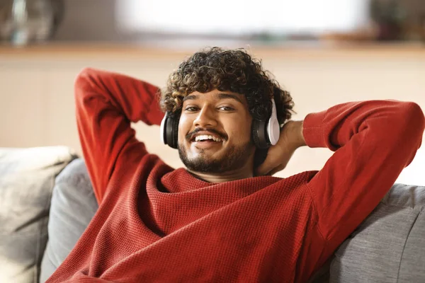 Happy Young Indian Guy Listening Music In Wireless At Home, Portrait Of Cheerful Handsome Eastern Man Relaxing On Couch With Hands Behind Head And Smiling At Camera, Enjoying Favorite Songs