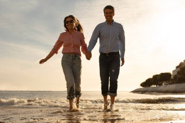 Romantik olgun çift, gün batımında plajda birlikte koşarken el ele tutuşuyor, orta yaşlı eşleri seviyor, yaz tatilinin tadını çıkarıyor, mutlu erkek ve kadın dışarıda çıkıyorlar, uzay kopyaları yapıyorlar.