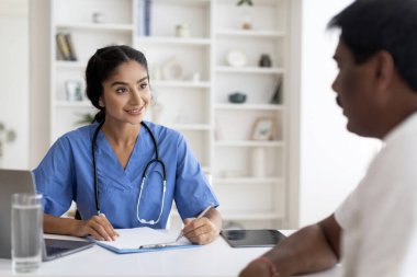 Ofisteki toplantı sırasında Hintli doktor kadınla sağlık sorunları yaşayan erkek hasta, terapist kadınla konuşan hasta adam, panoya not alan kadın doktor, seçici odaklanma.