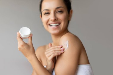 Beyaz banyo havlusuna sarılı nemlendirici krem kavanozu tutan neşeli genç bayan, duştan sonra omuz ve vücudu nemlendiriyor, gri stüdyo arka planında kameraya gülümsüyor. Seçici odak