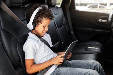 Otomobilde seyahat eden bir çocuk el bilgisayarı oyun tabletini paylaşıyor. Arabasının arka koltuğunda otururken dijital tablet kullanan kablosuz kulaklıklı Afro-Amerikalı çocuk.