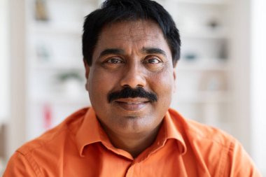 Bıyıklı, turuncu gömlekli, neşeli, orta yaşlı Hintli bir adamın yakın plan portresi. Evde ya da ofiste poz veriyor. Geçmişi bulanık. İnsanlar konseptle karşı karşıya