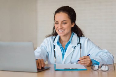 Profesyonel yardım konsepti. Beyaz üniformalı genç bayan doktor dizüstü bilgisayarla masada oturuyor, hastalara danışmanlık yaparken panoya yazı yazıyor, boşluğu kopyalıyor.