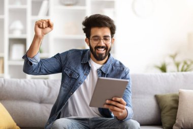 Neşeli Hintli adam evde dijital tablet bilgisayarla başarıyı kutluyor, rahat koltukta oturuyor ve heyecanla yumruğunu kaldırıyor, Şanslı adam çevrimiçi oyunda veya piyangoda bahis kazandı,