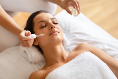 Yüz Spa 'sı. Genç bayan rahatlatıcı yüz masajı yaparken masör lüks spa salonunda cildini nemlendirirken havluya sarılı kadın cilt bakımı sırasında gözlerini kapatıyor.