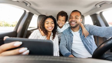Neşeli Afro-Amerikan ailesi otomobille seyahat ederken telefonda selfie çekiyor, kameraya gülümsüyor. Olumlu, havalı, prematüre siyah çocuk, anne ve babayı kucaklıyor, arabayla geziyorlar.