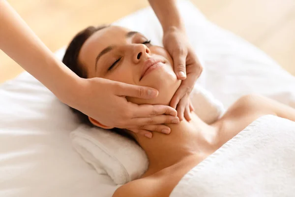 Millennial woman enjoying face building massage, lying with eyes closed while professional therapist making skin lift procedure indoor, high angle shot, closeup. Beauty treatment and spa
