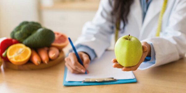 Female doctor making prescription or writing diet plan and holding green apple, sitting at desk with fresh fruits, cropped, panorama, closeup