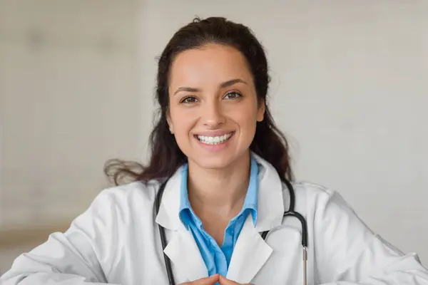 Professional help. Closeup portrait of cheerful female physician sitting at desk in clinic and smiling at camera. Lady doctor wearing white coat and stethoscope
