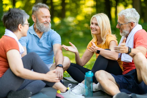 Group Of Happy Seniors Resting Together After Fitness Training In Park, Smiling Older Men And Women In Activewear Sitting On Fitness Mats, Chatting And Laughing, Relaxing After Workout Outside
