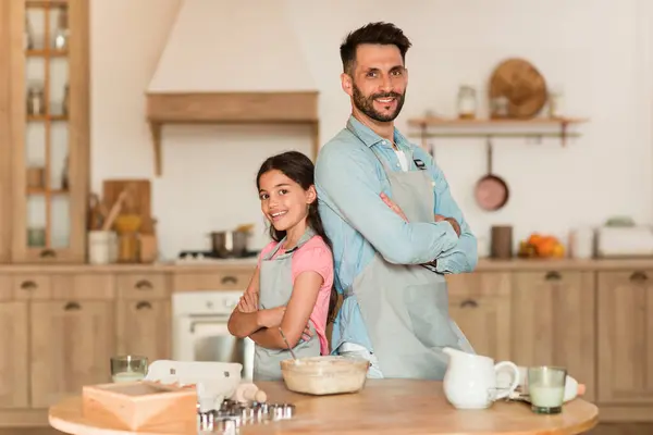 Family baking. Happy dad and his preteen daughter in aprons posing in kitchen with folded arms, standing back to back, ready for cooking pastry together