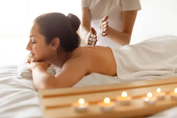 Spa And Beauty. Young Woman Having Relaxing Body Massage Session In Wellness Center Lying On Table Near Burning Candles Indoor, Professional Therapist Woman Massaging Her Back. Selective Focus