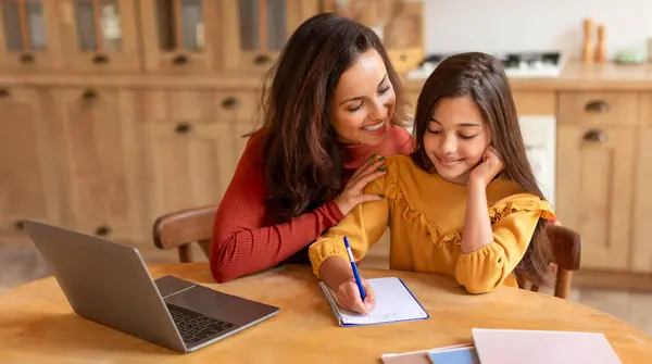 Smiling young mother helping her preteen daughter with homework at laptop, embracing smart kid girl as she writes, sitting together at kitchen table at home. Homeschooling concept. Panorama