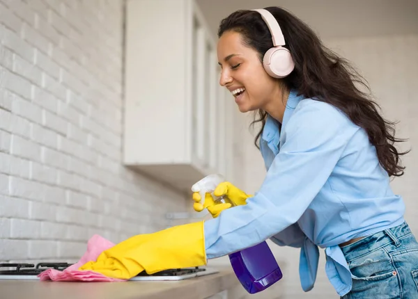 ヘッドフォンのハッピー主婦 キッチンカウンターを掃除し 音楽を聴く 家庭で整頓されたゴム製手袋を身に着けている女性 洗剤スプレーとラグを使用して 自由なスペース — ストック写真