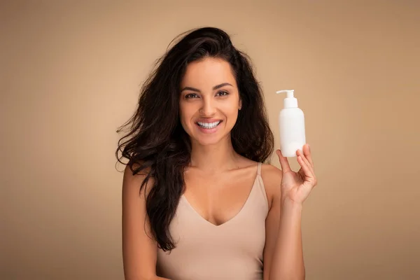 Attractive cheerful long-haired brunette eastern young woman wearing beige top holding small white bottle with beauty product, smiling, beige background. Cosmetics, hair treatment, body care routine