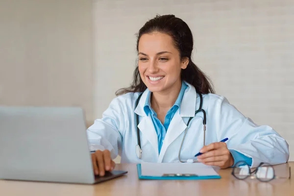 Professional help concept. Young female doctor in white uniform sitting at table with laptop, writing in clipboard while consulting patients online, copy space