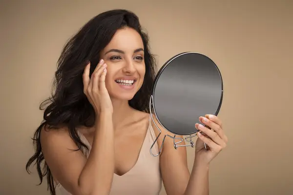 Anti-aging face care treatment, aesthetic procedures concept. Smiling attractive brunette long-haired young woman looking at mirror, touching face skin, beige studio background,, copy space