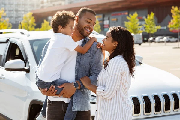 快乐的非洲裔美国家庭在他们全新的汽车旁边摆姿势 快乐可爱的黑人父亲 母亲和儿子站在漂亮的白色停车场旁 家庭汽车 — 图库照片