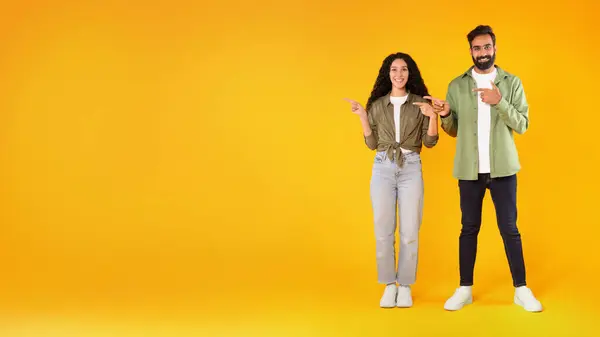 ナイス オファー 笑顔の若いアラブの配偶者は指を脇に置いて 黄色の背景にテキストの空白のスペースを示す両手でジェスチャーします カップルの広告の提供 パノラマの全長ショット — ストック写真