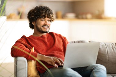 Güzel web sitesi. Evde dizüstü bilgisayar kullanan gülümseyen Hintli genç bir adamın portresi, bilgisayar üzerinde çalışan yakışıklı bir doğulu adam, oturma odasında koltukta otururken klavyede daktilo yazıyor, boşluğu kopyalıyor.