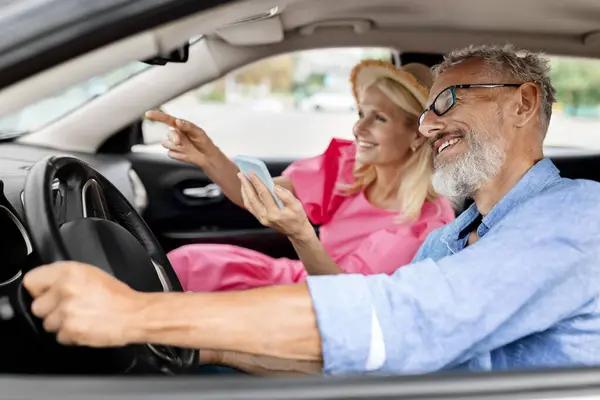 Happy elderly man and woman having car trip together, using cell phone and smiling, copy space. Cheerful retired couple travelers enjoying journey by cozy auto, tracking their way on smartphone gps