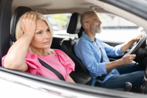 Upset attractive blonde senior woman in pink dress sitting next to her husband driving auto, touching head, look through window, feeling down after quarrel with spouse. Family fight during car ride