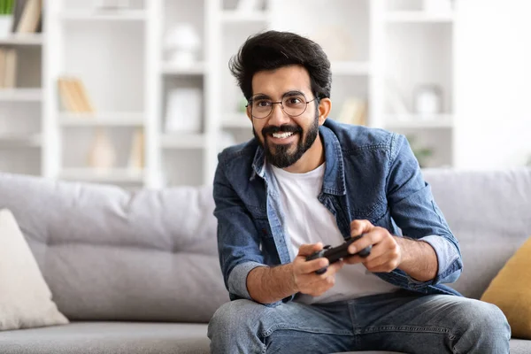 Portrait of happy excited young indian guy playing video games at home, handsome eastern man holding console joystick and smiling, gamer guy sitting on the couch in living room, copy space