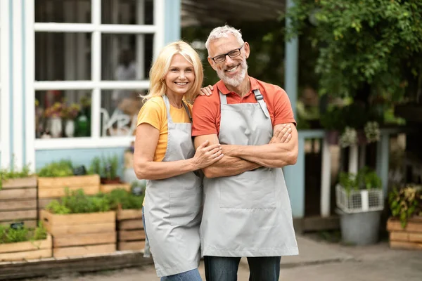Portrait Of Happy Mature Farmers Couple Standing Outdoors Near Their Greenhouse, Cheerful Senior Man And Woman Small Business Owners Wearing Aprons Posing Outside, Looking And Smiling At Camera