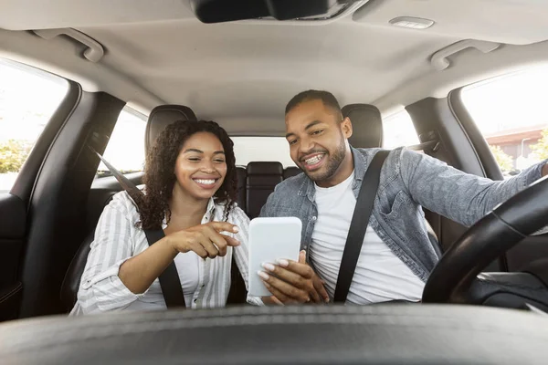 Millennial black man and woman have car trip together, using cell phone. Happy african american couple travelers enjoying journey by cozy auto, track their way on smartphone, booking hotel
