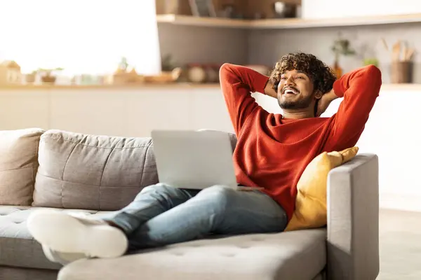 Lazy Day. Smiling Young Indian Man Relaxing At Home With Laptop Computer, Handsome Happy Eastern Guy Leaning Back On Couch With Hands Behind Head, Resting In Living Room Interior, Copy Space