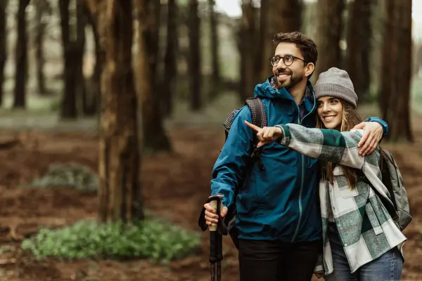 Smiling young caucasian lady in jacket pointing finger at empty space for man, enjoy adventure, travel in cold forest in autumn. Vacation together, active lifestyle, free time at weekend and walk
