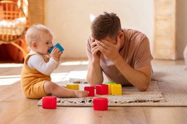 Bebekli yorgun bir baba evde oyuncak oynuyor, depresif bir adam oturma odasında dinlenirken çocuğuna bakıyor, baba olmaktan rahatsız oluyor, ebeveyn yorgunluğu çekiyor.
