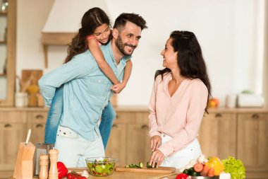 Annesi vejetaryen salatası pişirirken, sohbet ederken ve mutfak içindeyken, mutlu bir baba kızını sırtında taşır. Birliktelik kavramı.