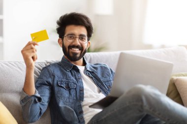 İnternetten alışveriş. Gülümseyen Hintli adam dizüstü bilgisayar tutuyor ve evde koltukta otururken kredi kartı gösteriyor, yakışıklı doğu çocuğu kolay internet ödemeleri veya para transferleri, bedava alan öneriyor.